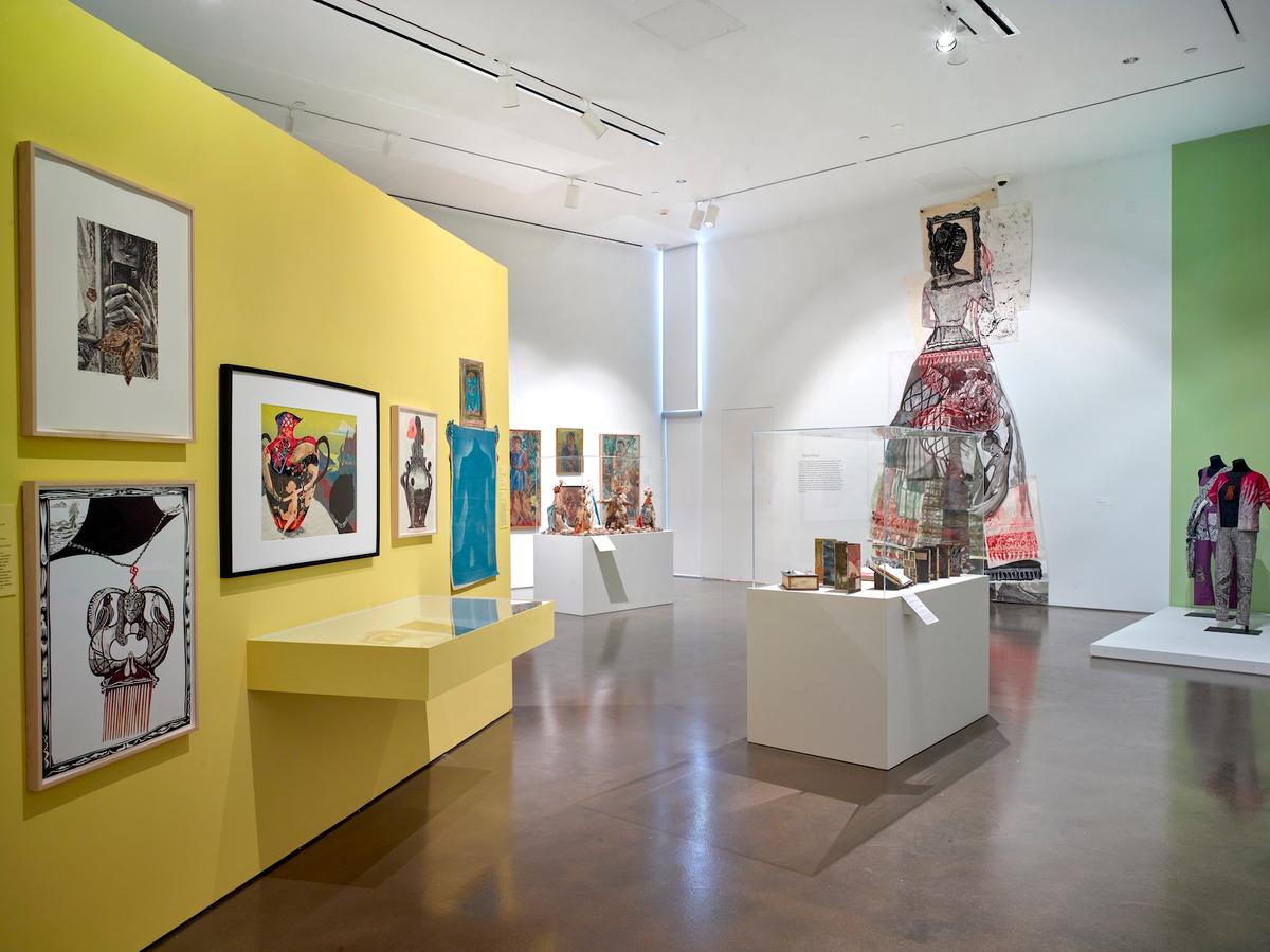 Bright yellow wall with framed illustrations, adjacent to sculptures, mannequins, and textile artworks in the gallery space.