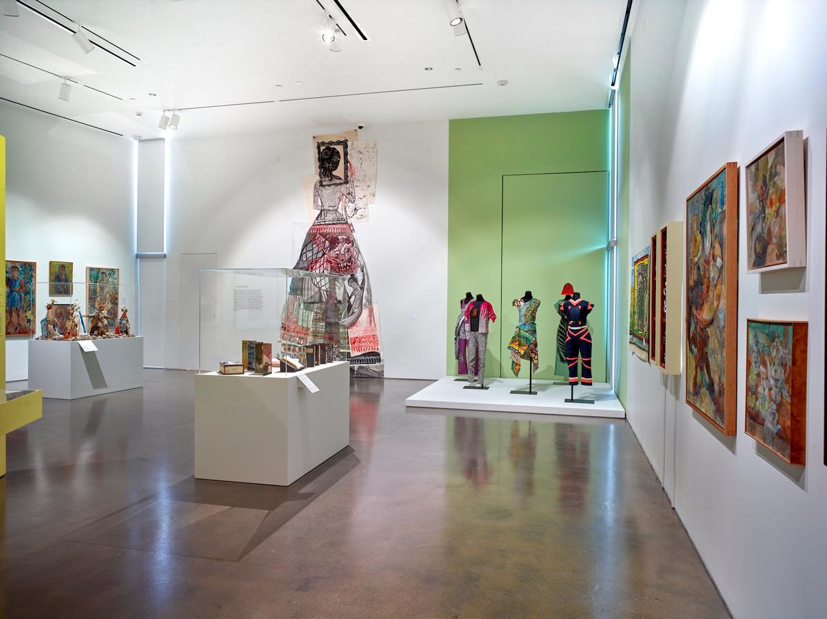Gallery view featuring colorful garments on mannequins, vibrant paintings on the walls, and a large dress form under spotlight.