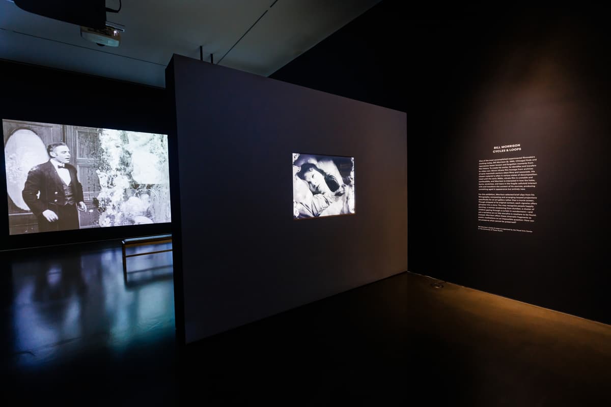 Two black-and-white film projections in a gallery; in the background on the left side a screen shows a suited man, on the right and in front, a screen shows a woman lying in bed, with introductory wall text nearby.