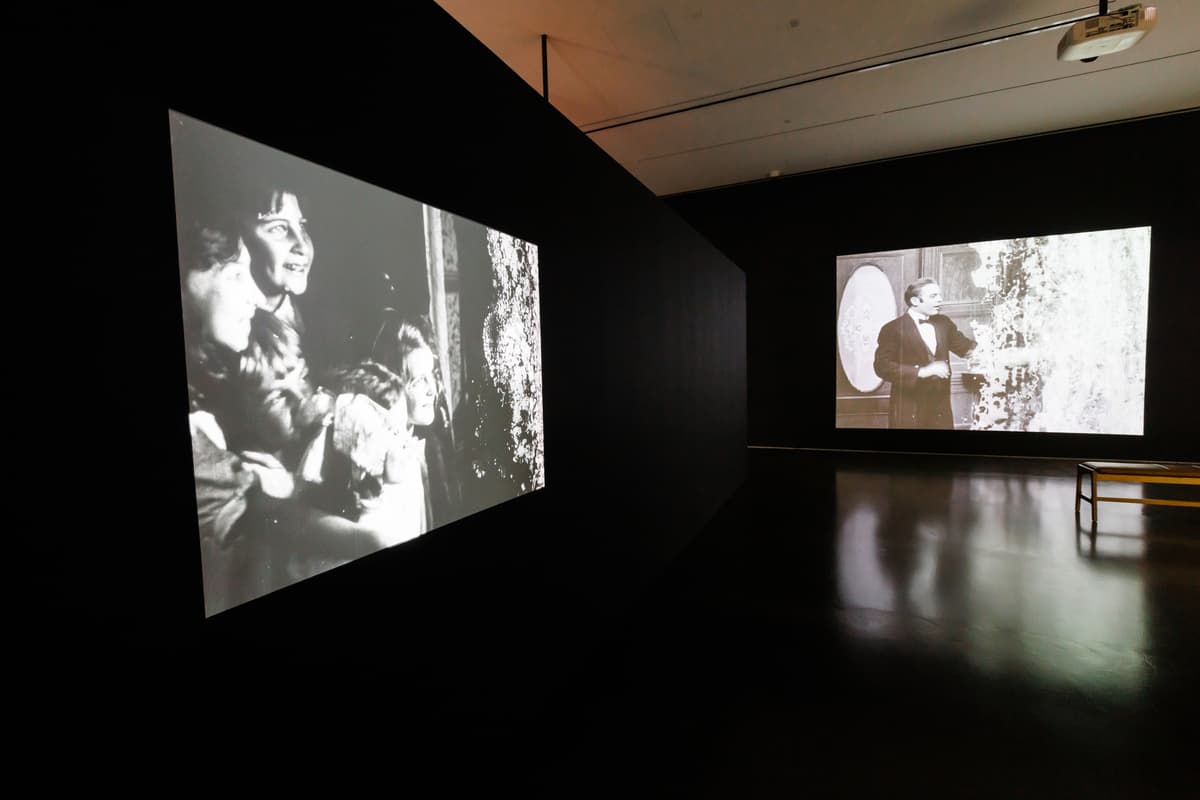 Two black-and-white film projections on adjacent walls in a dark gallery; the one on the left wall shows a group of children smiling, and the one on the right wall shows a man in a suit.