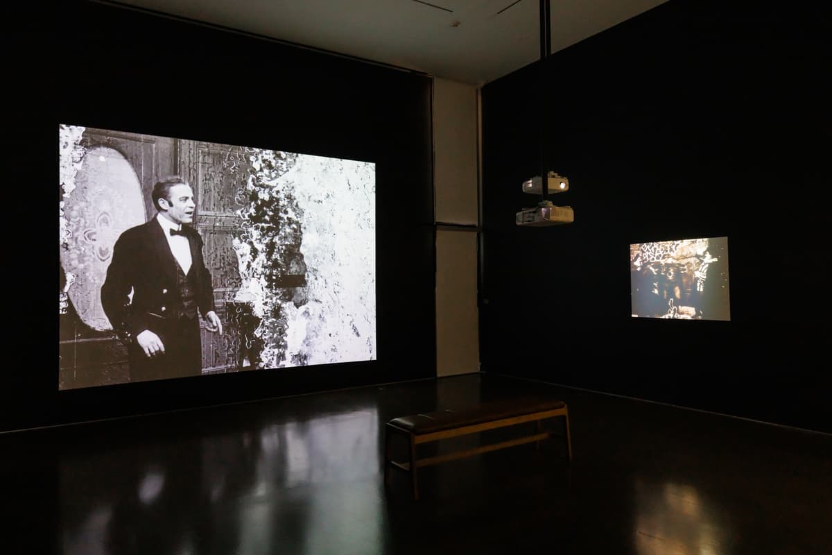 Projected black-and-white film of a man in a suit on the left wall, with a smaller projection and bench visible in a dark gallery space.