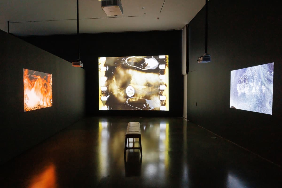 Installation view of three video projections displayed on gallery walls with a central wooden bench for viewers.