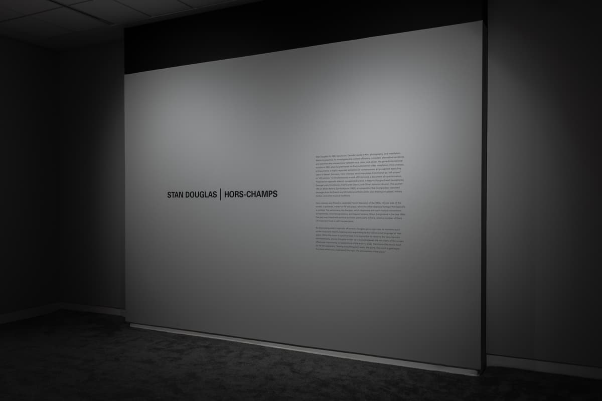 Gallery corner with a wall reading “STAN DOUGLAS | HORS-CHAMPS” and an exhibition text panel to the right