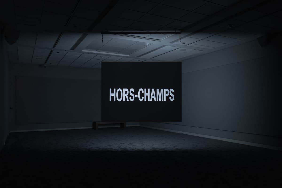 Dark gallery space with a projected black screen displaying the text “HORS-CHAMPS” in large white letters