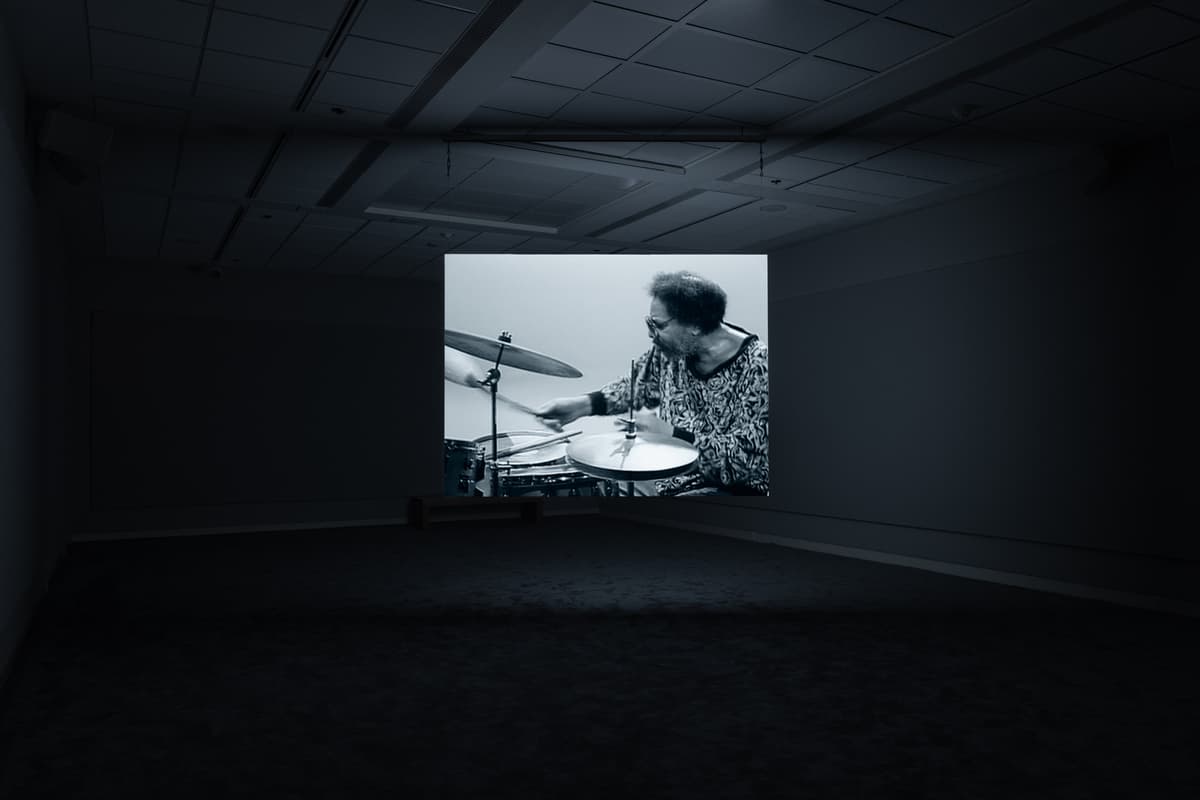 Dark gallery space with a projected black-and-white image of a drummer playing a drum kit