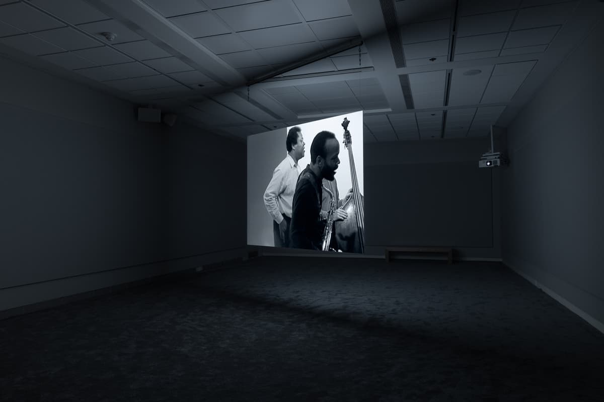 Dark gallery space with a projected black-and-white image of two men, one playing an upright bass while the other looks on