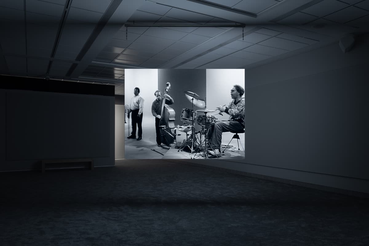 Dark gallery space with a projected black-and-white image of a jazz band, showing musicians with a bass, drums, and saxophone