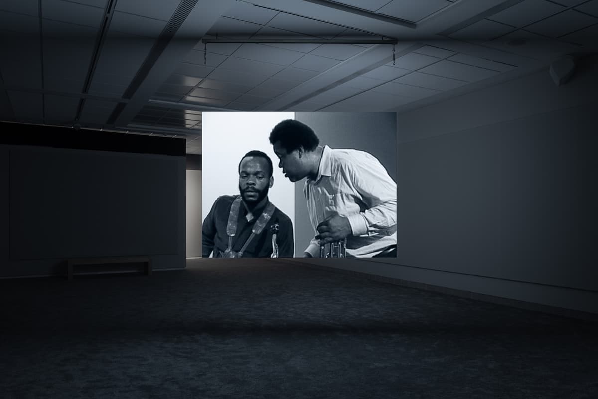 Dark gallery space with a projected black-and-white image showing two men in conversation, one seated and the other leaning in