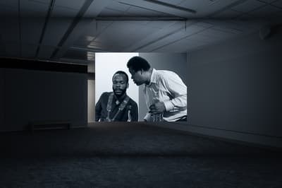 Dark gallery space with a projected black-and-white image showing two men in conversation, one seated and the other leaning in