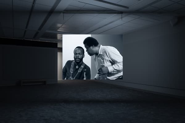 Dark gallery space with a projected black-and-white image showing two men in conversation, one seated and the other leaning in