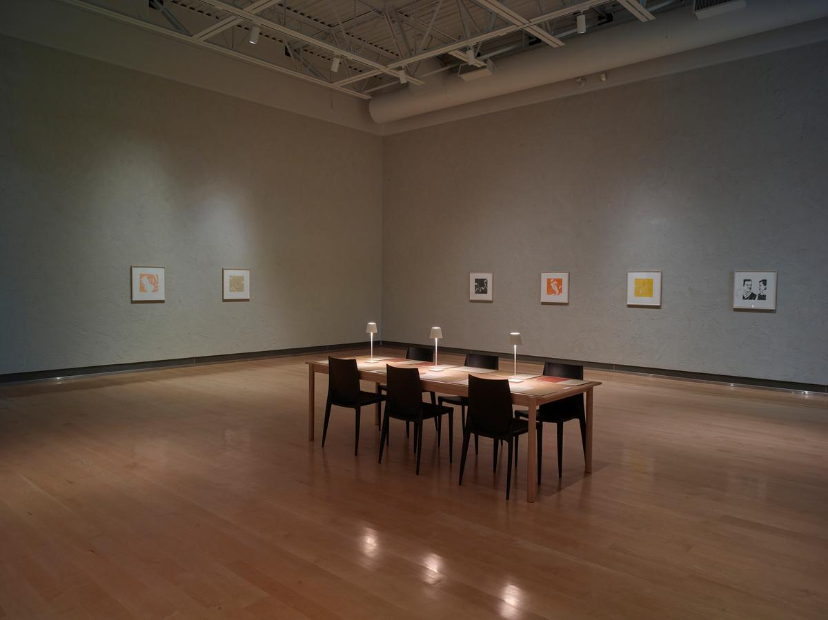 Reading table with books and chairs placed centrally in a gallery space, surrounded by small framed works hung in a linear arrangement on textured gray walls.