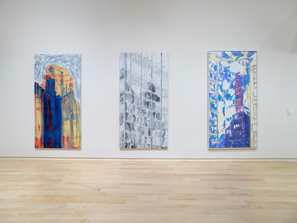 Three large vertical cityscape paintings in vibrant colors hanging on a white wall with a light wooden floor in a museum gallery.
