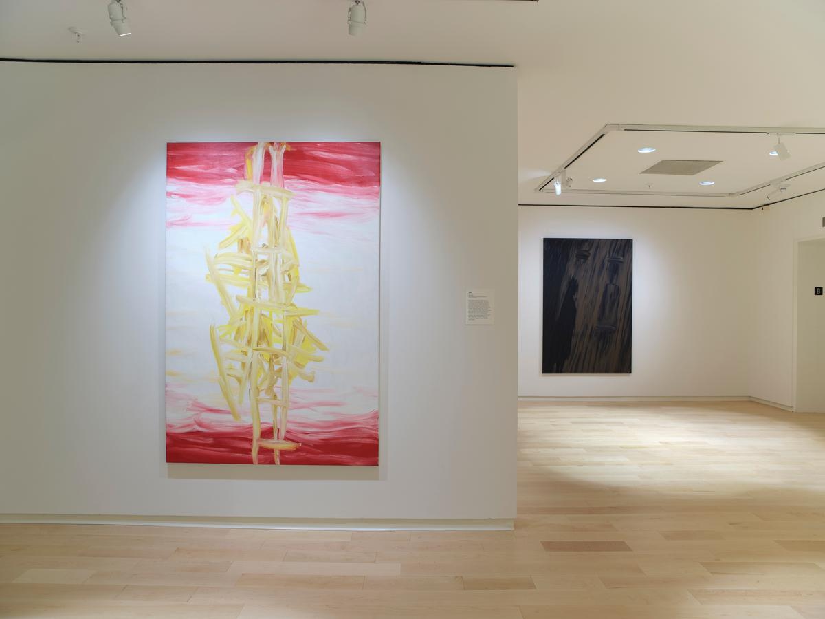 Large abstract painting with yellow vertical forms and red cloud-like edges installed beside a gallery entryway, with a dark abstract painting visible in the adjacent room.