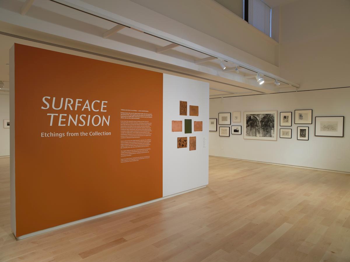 Orange title wall reading “Surface Tension” with curatorial text, adjacent to white wall featuring small abstract etchings and framed prints