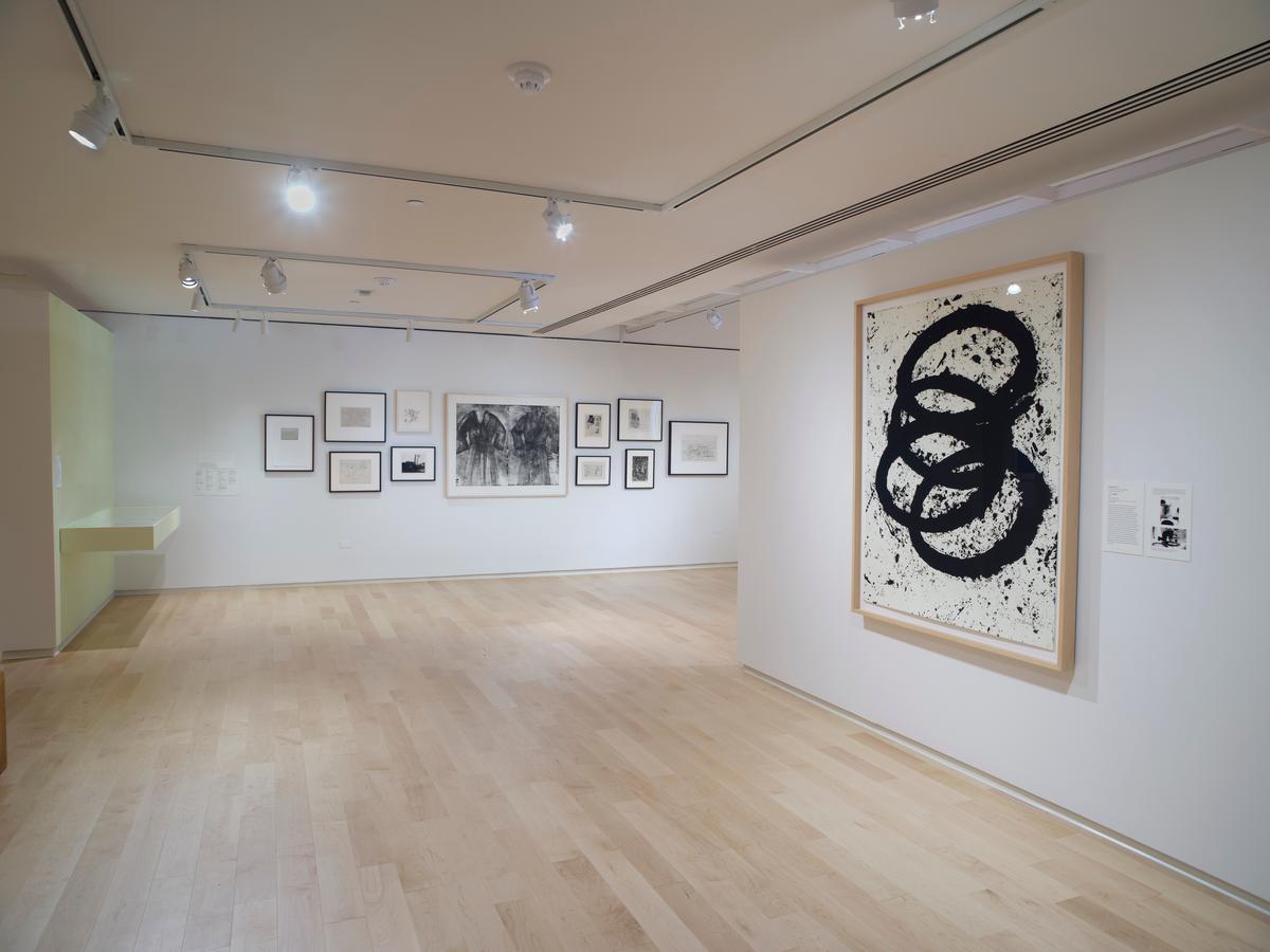 Large gestural black ink print on right wall with a salon-style arrangement of etchings on back wall in white-walled gallery