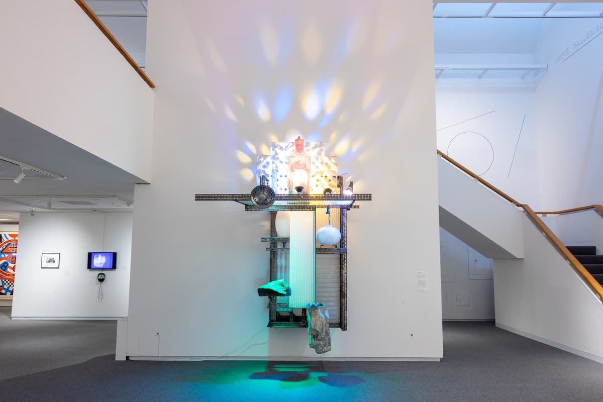 Mixed-media wall sculpture illuminated with multicolored light projections positioned near a stairwell.