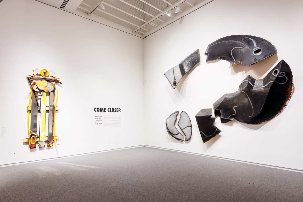 Two sculptural wall works displayed near “Come Closer” block lettering; one work is composed of dark curved metal fragments and the other features yellow and red structural elements