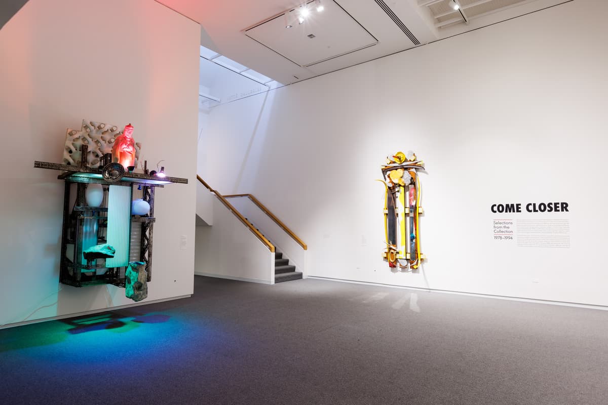 Two wall mounted sculptures on adjacent walls next to vinyl block lettering reading "Come Closer," the exhibition title, with introductory text.