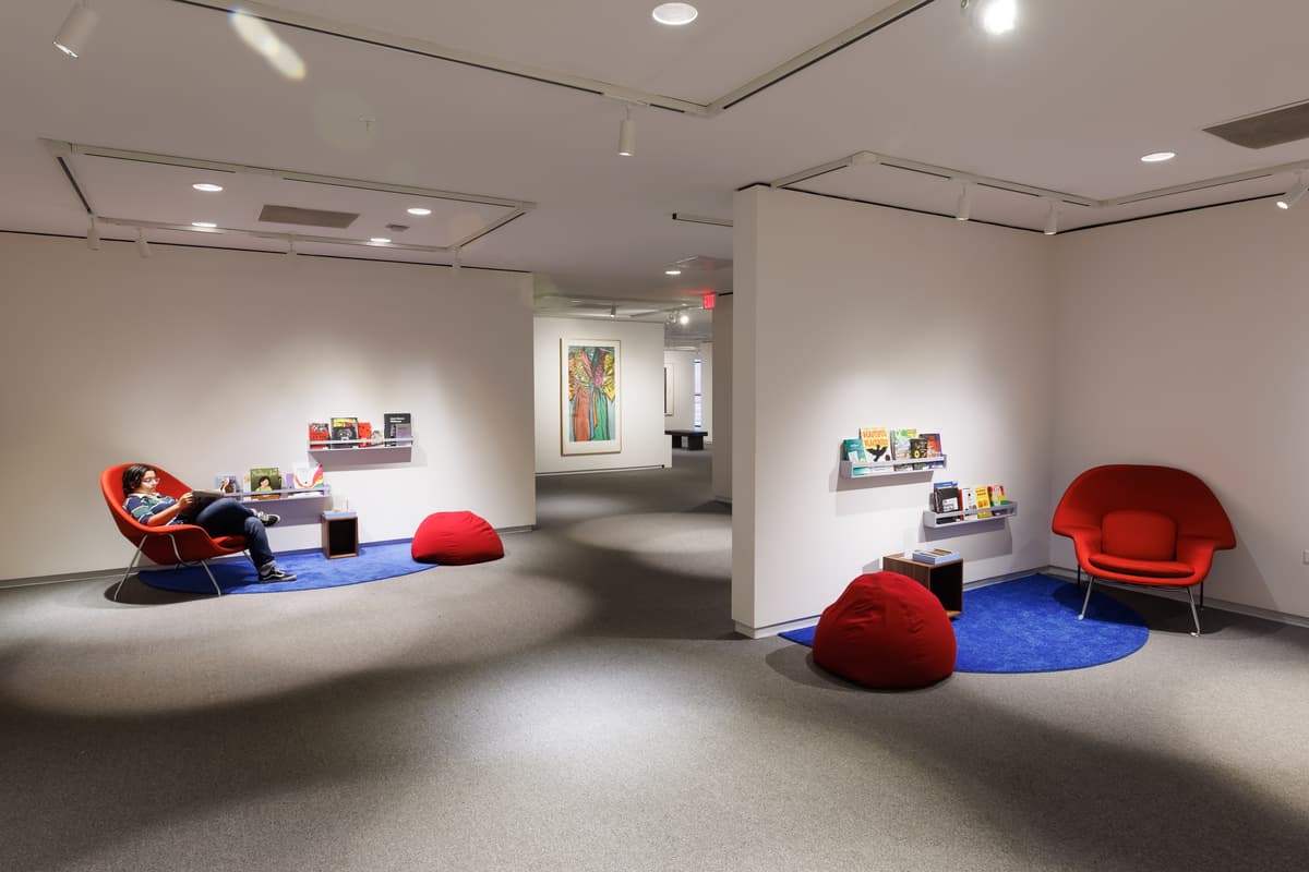 Two reading nooks each with a red armchair and red beanbag on a blue area rug; a pair of floating shelves and side table hold books for visitors.