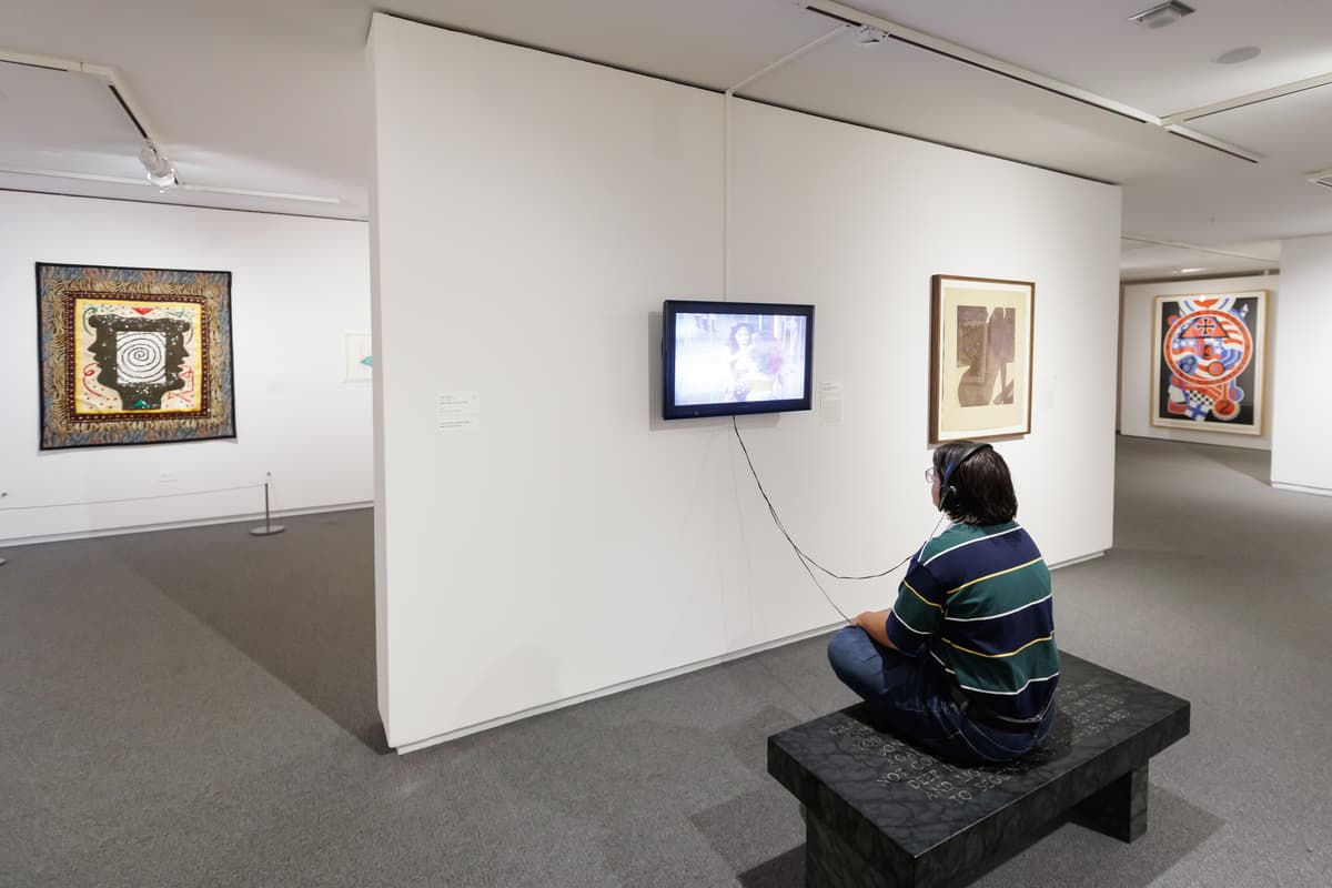 Person seated on a bench watching a wall-mounted video screen with headphones, surrounded by colorful and abstract artworks.