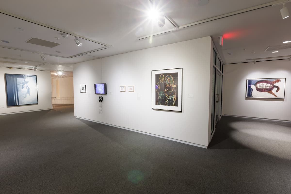 Various framed artworks and a wall-mounted video screen in a white-walled gallery space.
