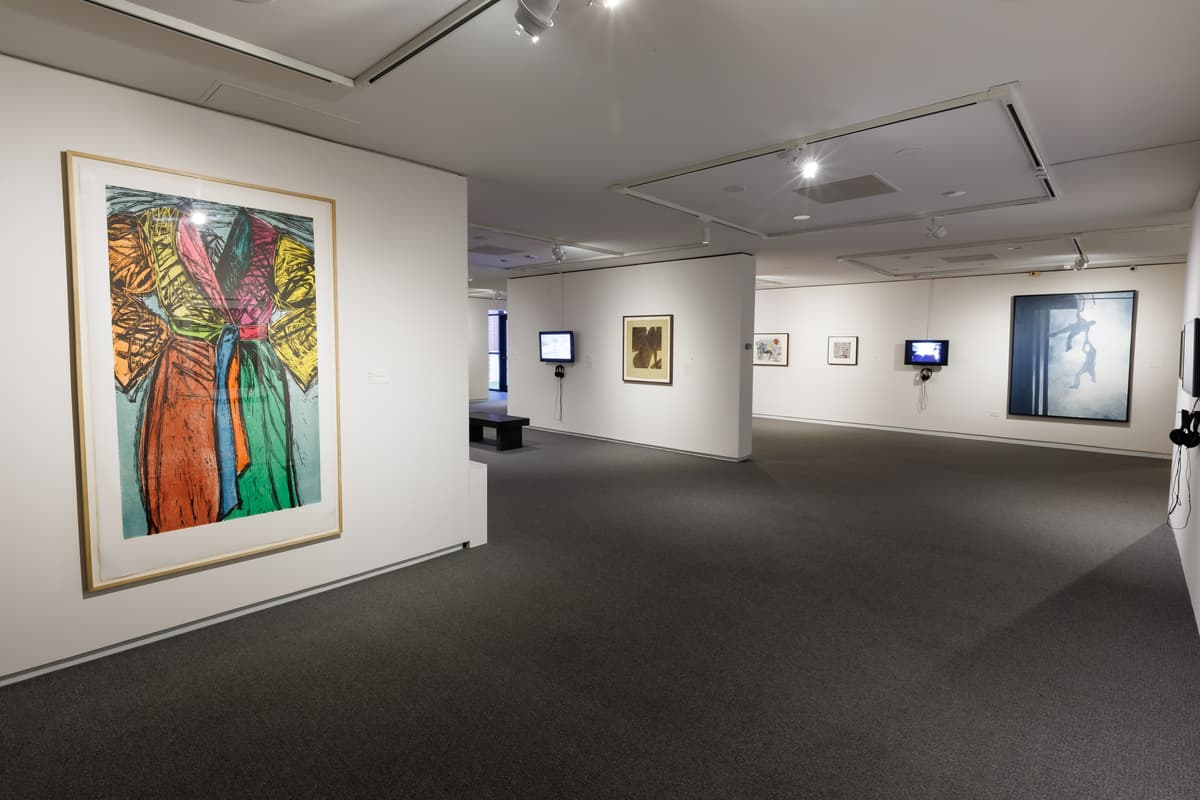 Large colorful abstract print on the left wall depicting a robe with additional framed works and wall-mounted video screens visible throughout the gallery.