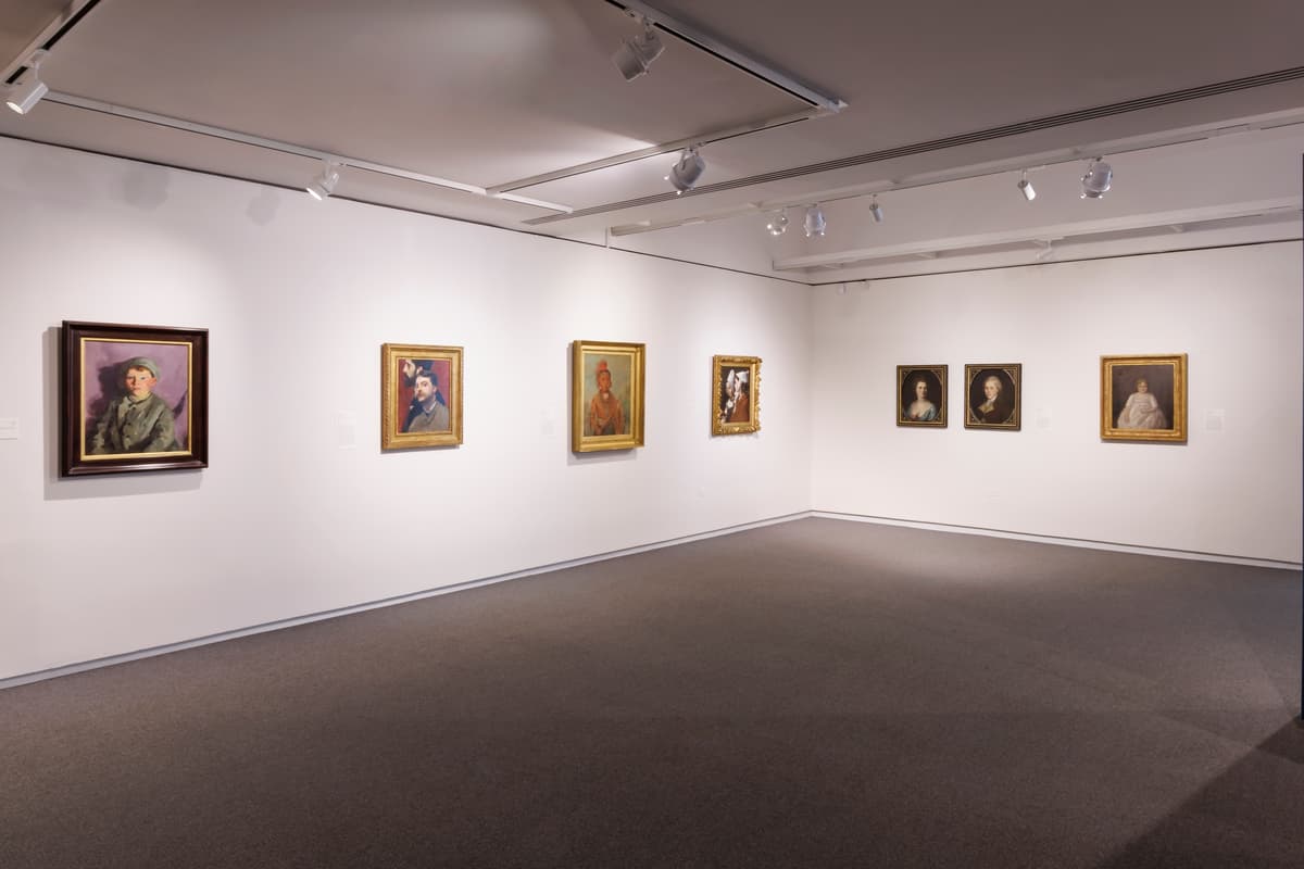 A row of seven portrait paintings on two adjoining white walls.