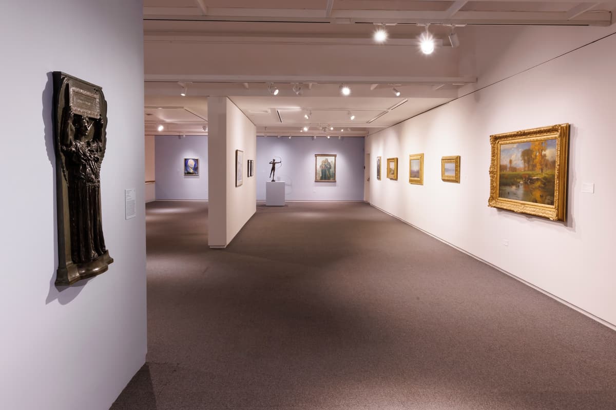 Gallery view with a bronze relief sculpture on the left, landscape paintings on the right wall, and a bronze figurative sculpture in the background.
