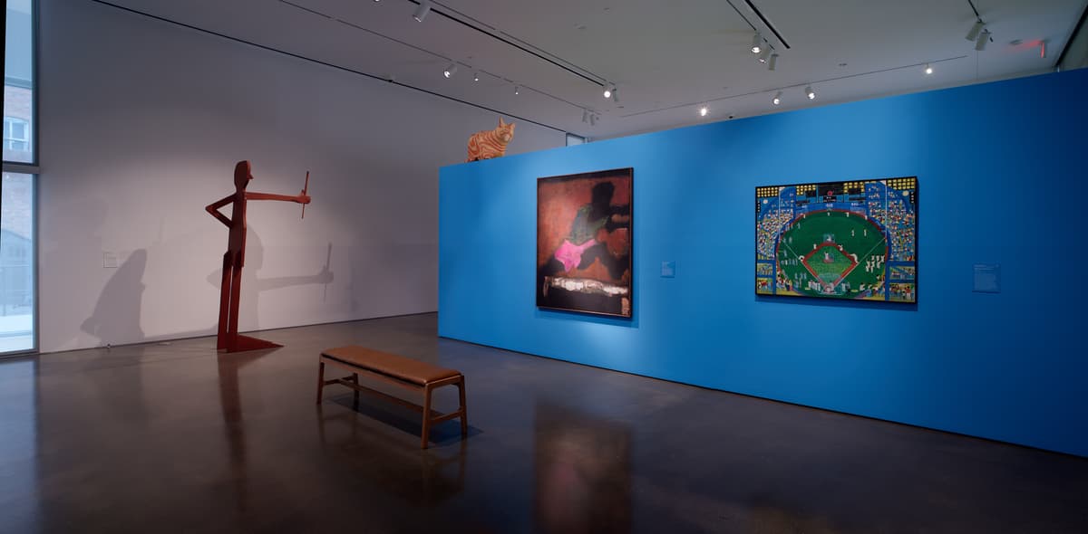 Sculpture of a figure holding a baton next to two paintings on a blue wall and a wooden bench.