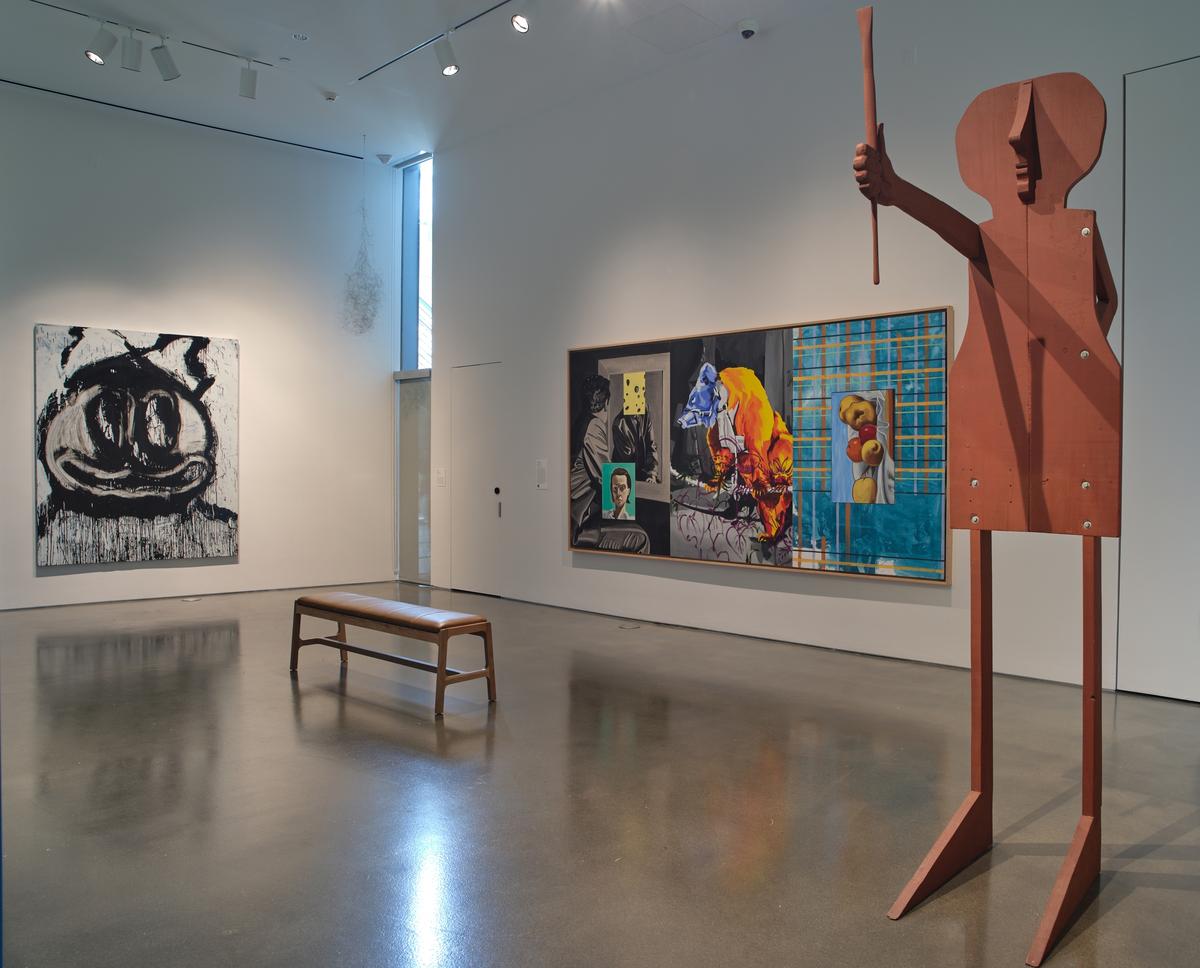Two paintings hanging behind a bench and a tall wooden sculpture figure holding a baton.