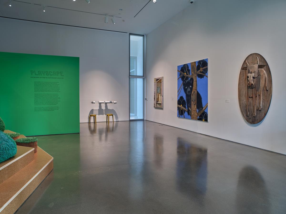 A green wall with exhibition text next to wall-mounted artworks including a large blue-toned painting and a wooden oval relief, each depicting animals.