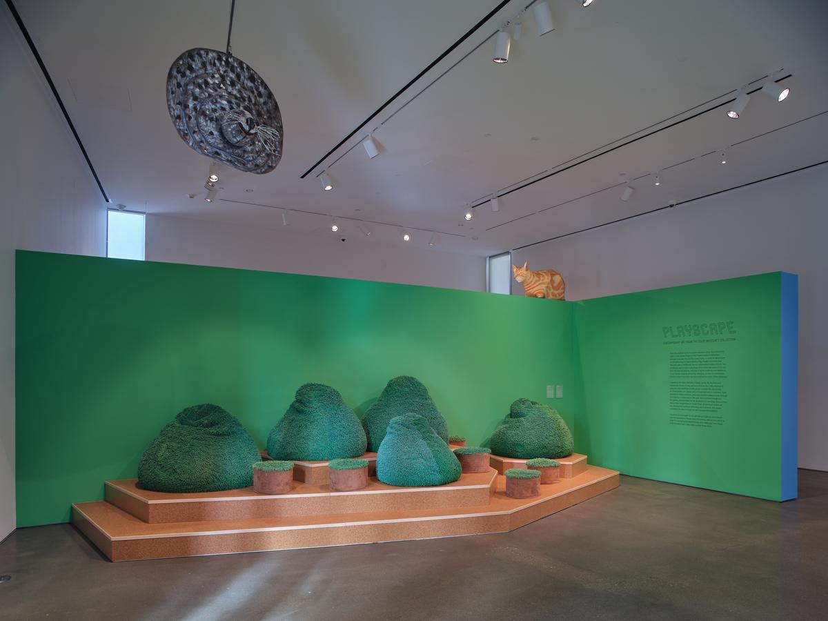 Rounded sculptural forms resembling hedges on a terraced platform against a green wall. Above, a sculpture of a cat rests on the wall and a second sculpture depicting the face of a seal is suspended from the ceiling.