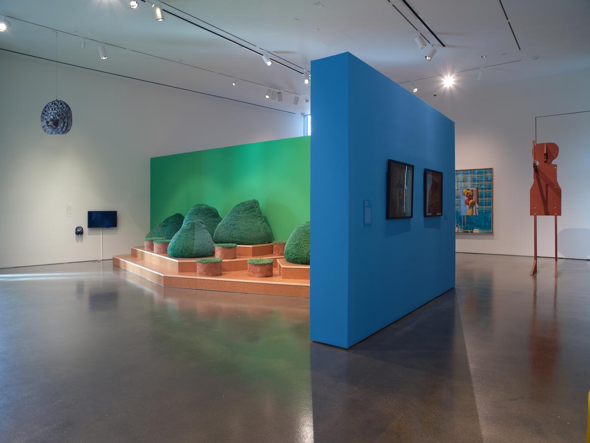 Gallery space with wooden stepped platform with rounded hedge-like sculptures against a green wall, a blue wall with mounted screens, and a tall abstract figure in the background.