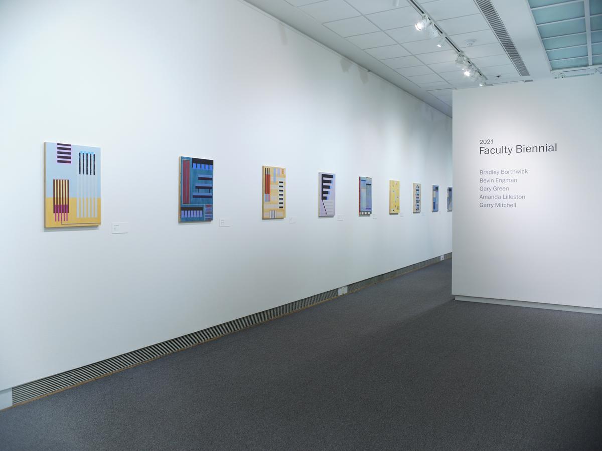 Colorful geometric prints in a row on a long white gallery wall with exhibition title text on an adjacent wall