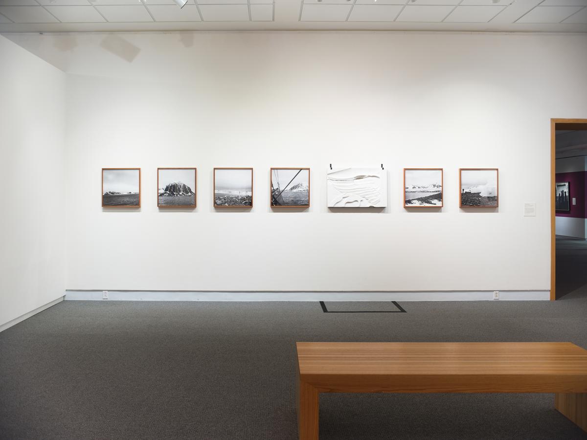 Seven framed black-and-white landscape photographs in a row on a white gallery wall with a wooden bench in front