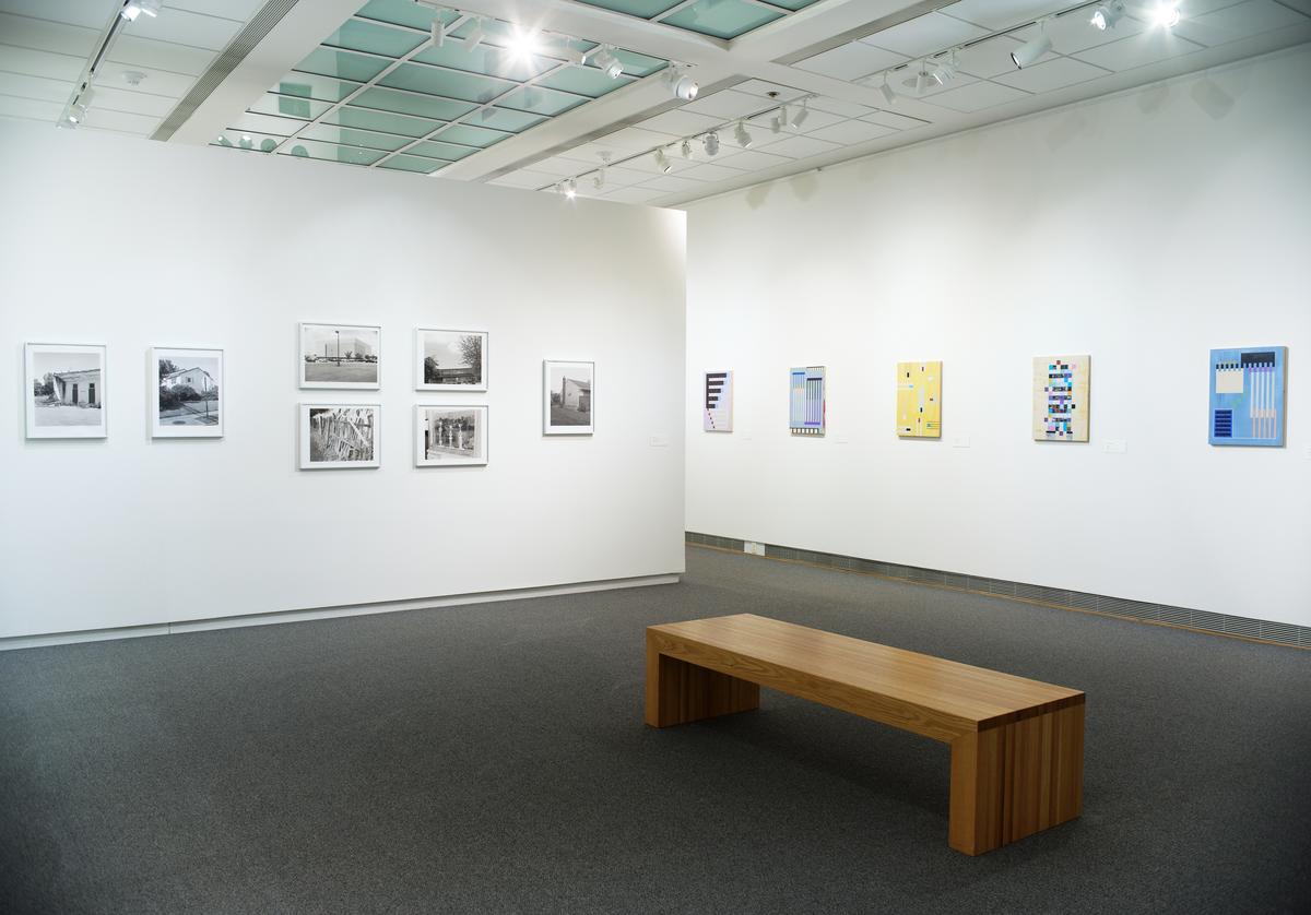 Gallery view with black-and-white photographs on one wall and colorful geometric prints on another, with a wooden bench in the center