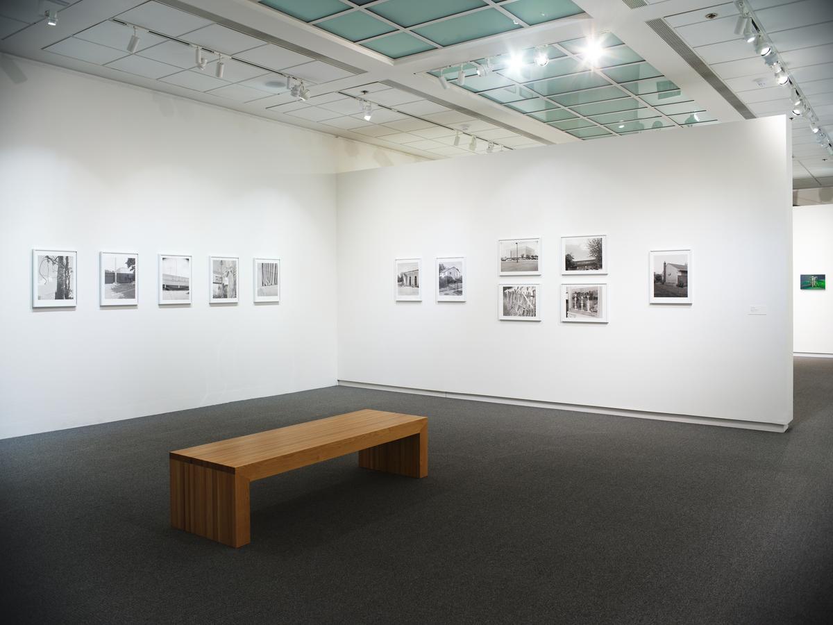 Black-and-white photographs arranged on two adjacent white gallery walls with a wooden bench in the center