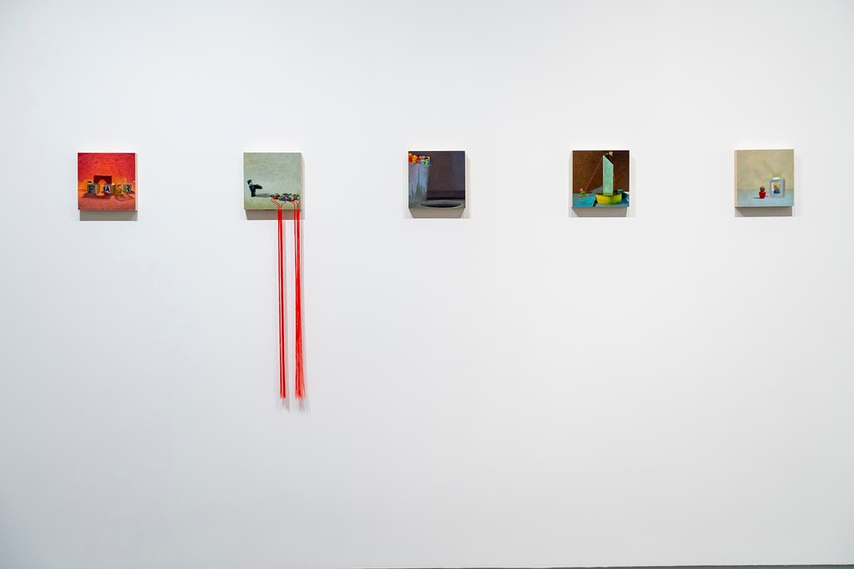 Five small colorful paintings mounted on a white wall, one featuring red threads hanging down from the canvas.