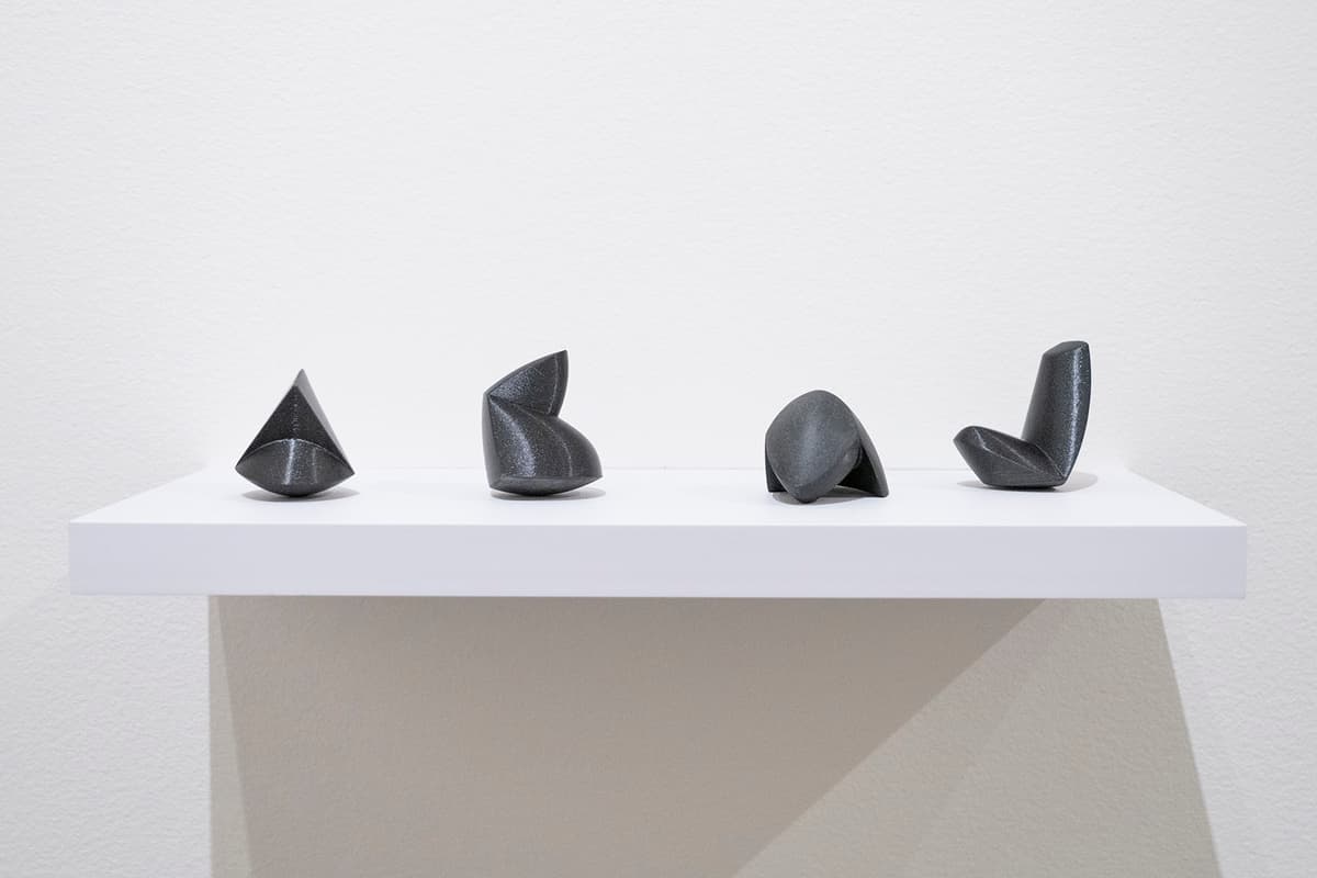 Four small, dark abstract sculptures displayed on a minimalist white shelf against a white wall.
