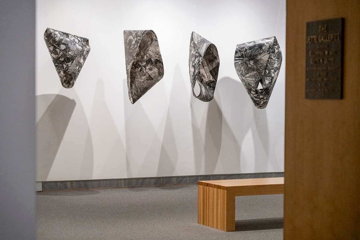 Four suspended abstract forms with marbled black and white surfaces, casting shadows on a white gallery wall, with a wooden bench in the foreground.