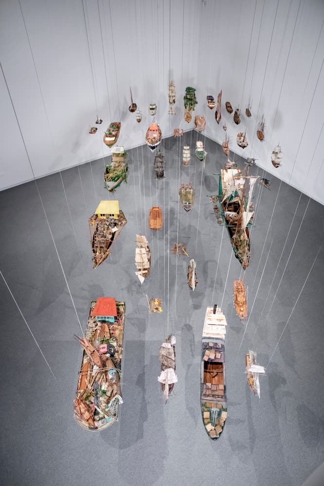 Overhead view of suspended boat sculptures arranged in rows, casting shadows on the gallery floor