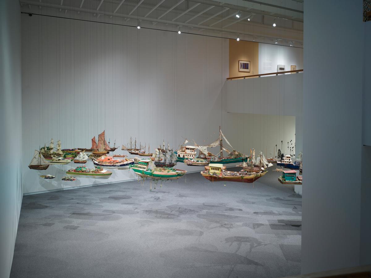 Colorful model ships suspended mid-air across a white-walled gallery with gray carpet