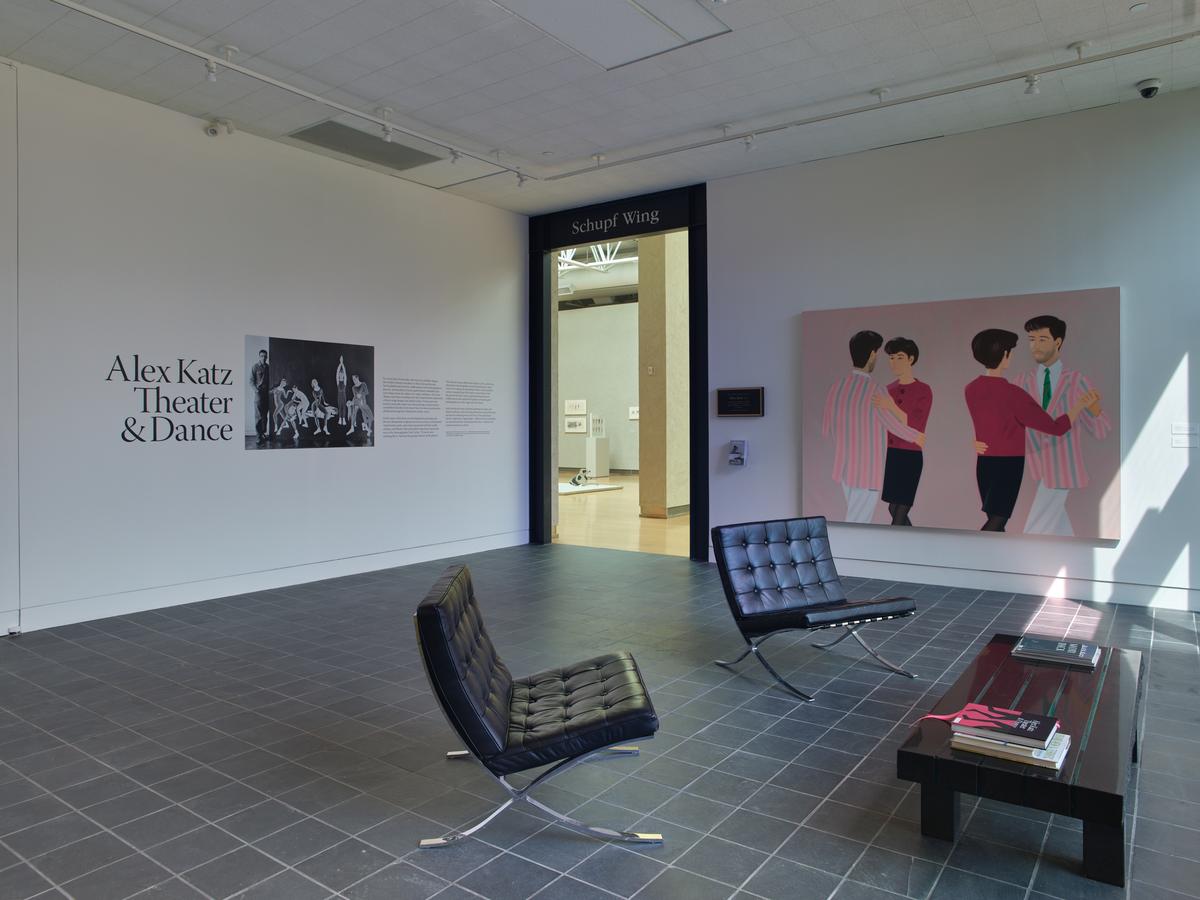 Lobby with coffee table and two chairs positioned in front of a wall with exhibition introduction text with title "Alex Katz Theater & Dance, a black-and-white photograph of the artist with an artwork, and several paragraphs of text." On the adjacent wall, a doorway labeled "Schupf Wing" and a painting with two figures dancing.