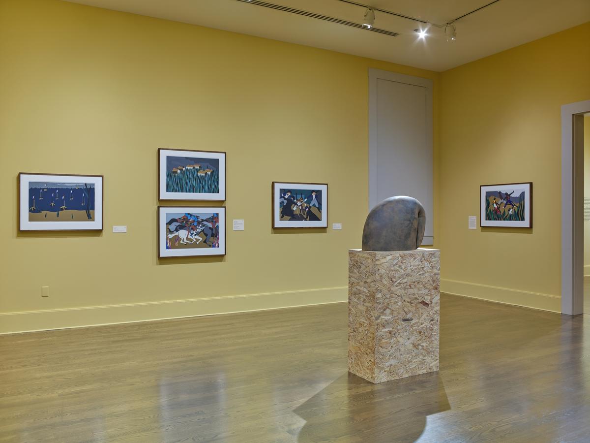 Framed narrative paintings on a yellow gallery wall with a smooth stone sculpture on a wooden pedestal in the foreground