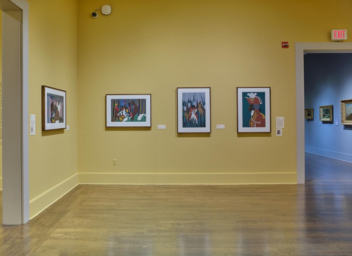 Framed narrative paintings on a yellow gallery wall near a doorway leading to another exhibition space