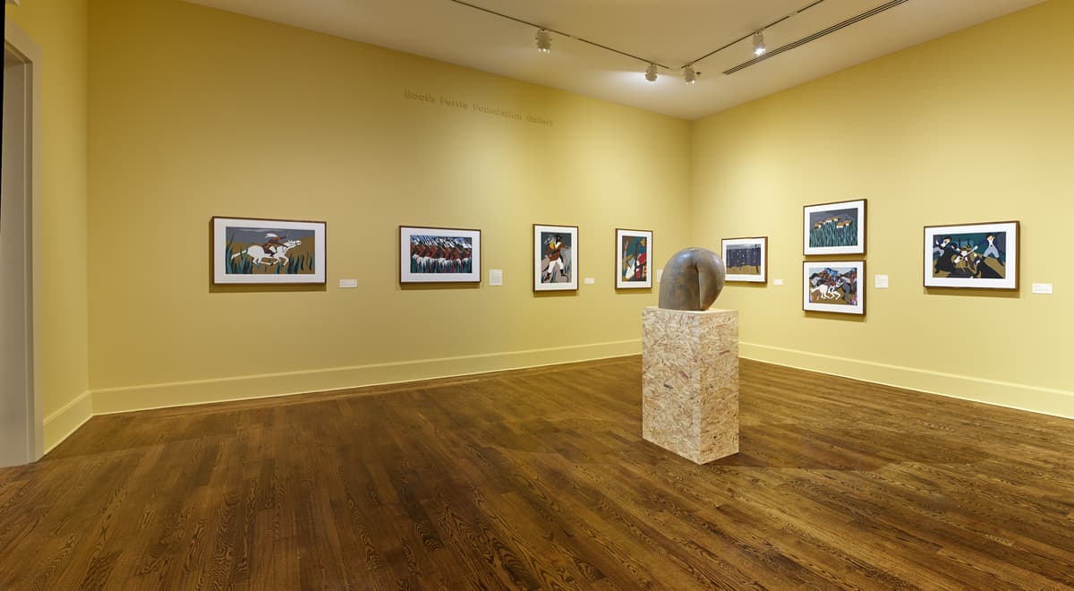 Panoramic view of yellow-walled gallery with framed narrative paintings and a stone sculpture on a pedestal