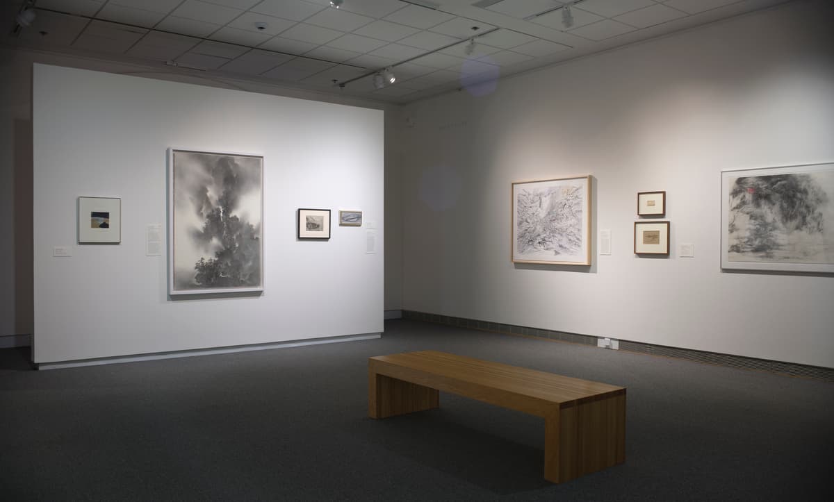 Framed abstract and landscape artworks on two white gallery walls with a wooden bench in the center