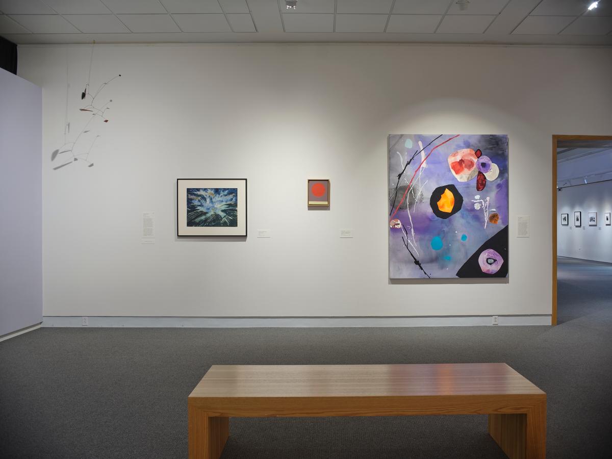 Gallery wall with three colorful artworks, a white hanging mobile, and a wooden bench in the foreground