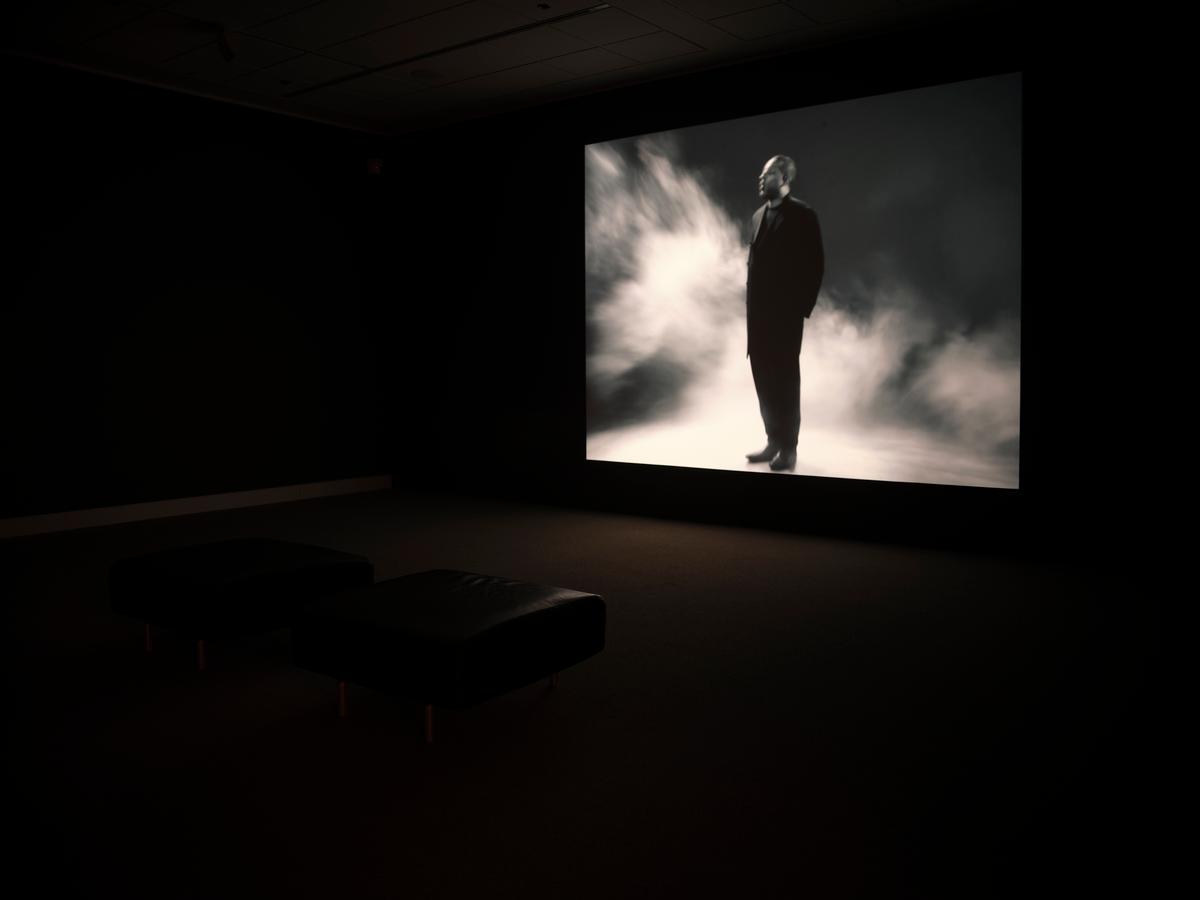 Dark gallery room with video projection of a figure in a suit standing in fog, with black benches in front