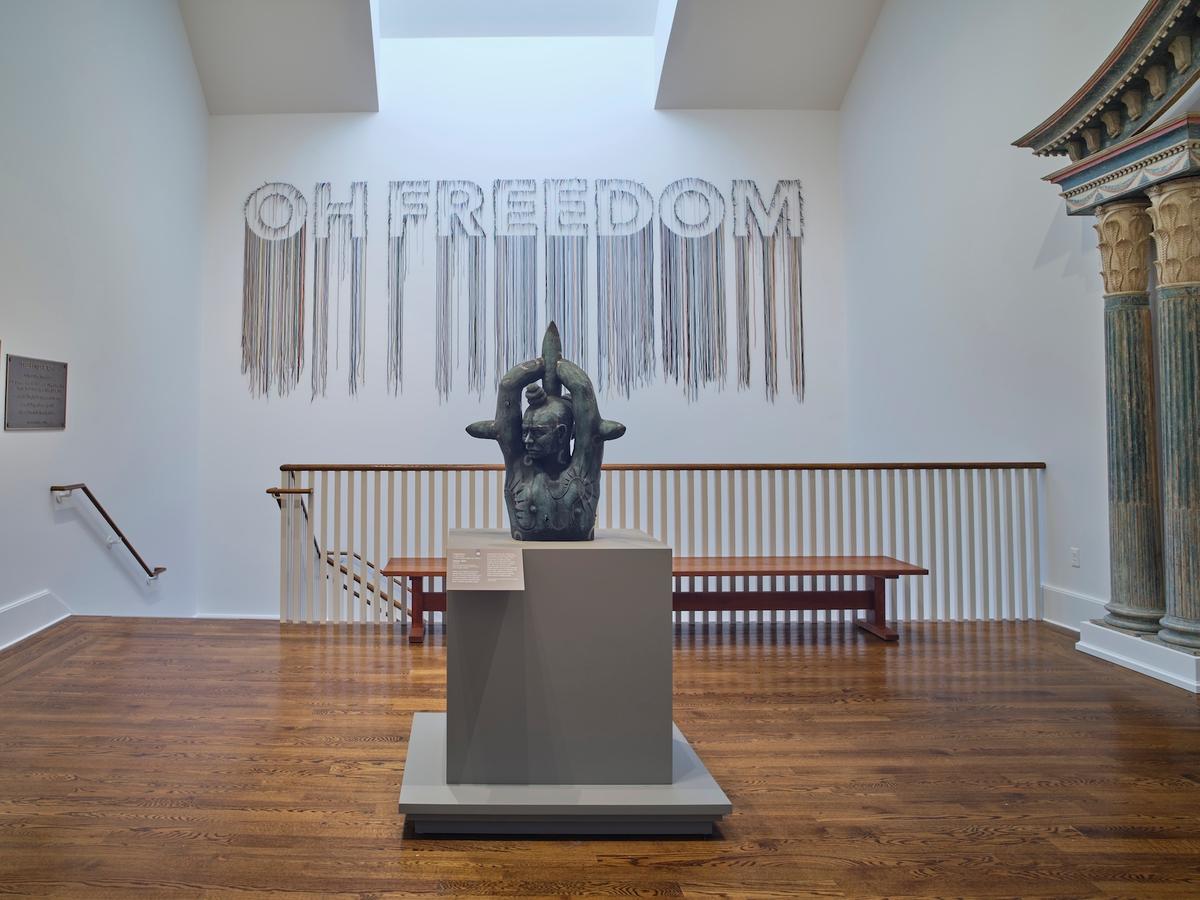 Sculpture on pedestal and an installation reading “OH FREEDOM” on the back wall spelled with hanging strands, positioned in a stairwell space.